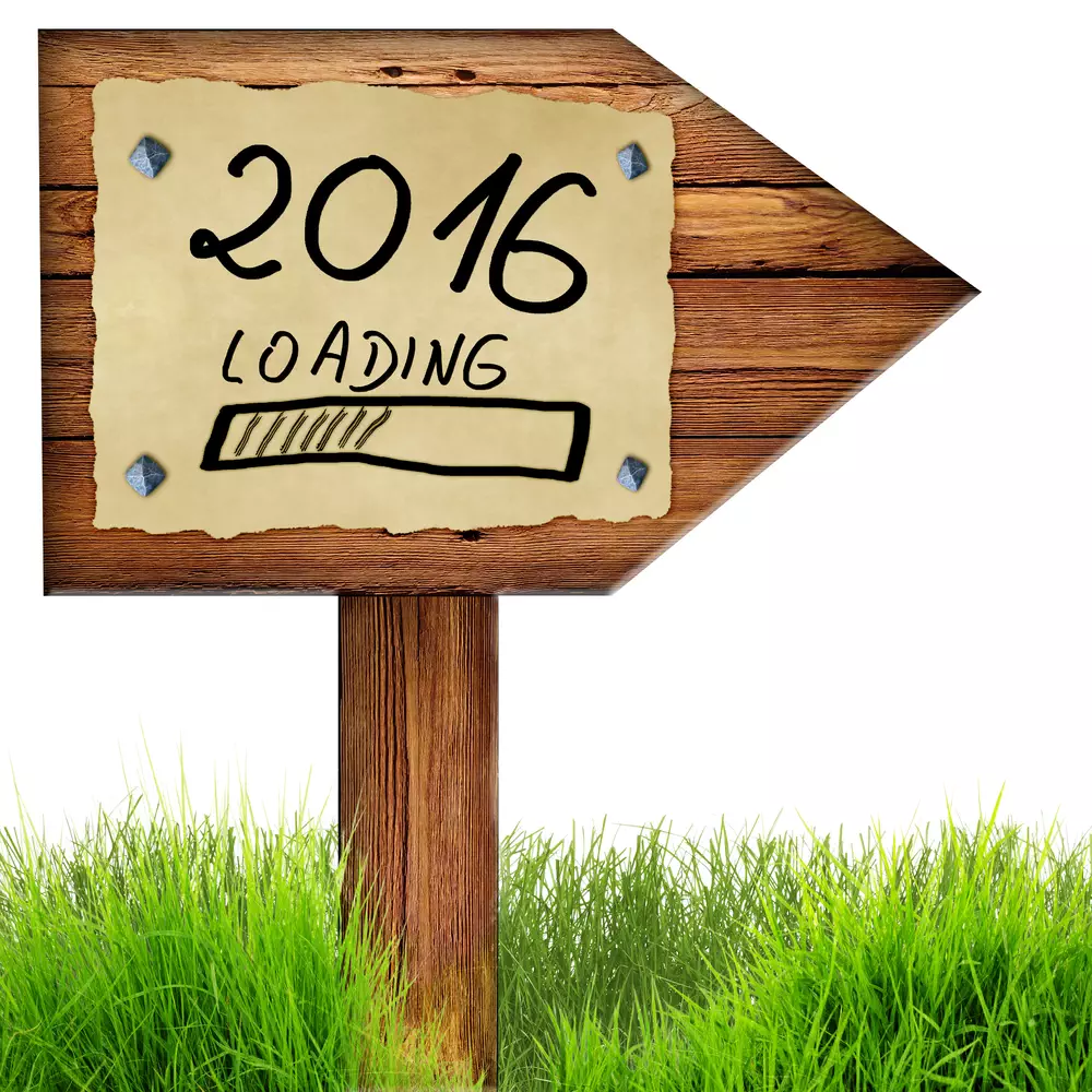 Wood arrow sign with 2016 loading handwritten on old page of paper nailed to planks, green grass around, isolated on a white background.
