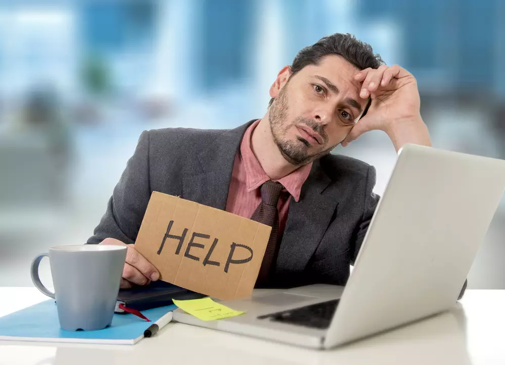 young businessman at office desk working on computer laptop asking for help holding cardboard sign looking desperate and depressed in business stress overwhelmed and overwork concept