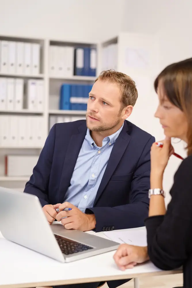 Serious young businessman sitting listening and watching something off screen as he sits sharing a laptop with a female colleague