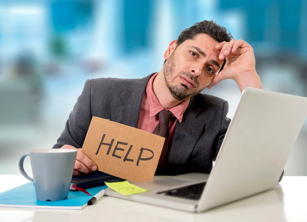 young businessman at office desk working on computer laptop asking for help holding cardboard sign looking desperate and depressed in business stress overwhelmed and overwork concept